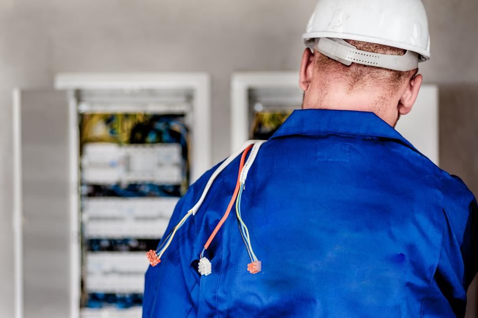 Een elektricien in blauwe werkkleding, van achteren, voor twee open zekeringkasten.