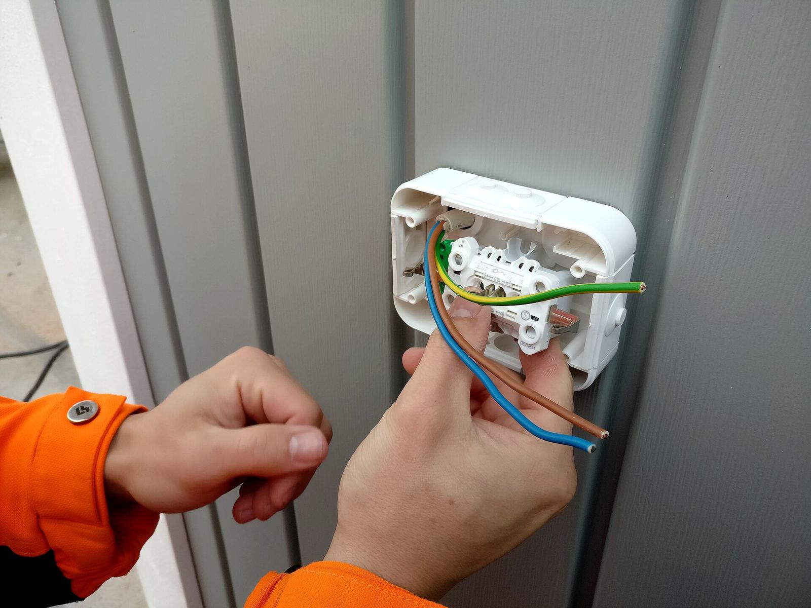 Twee handen sluiten een opbouwstopcontact aan: een bruine, een blauwe en een geel-groene draad.