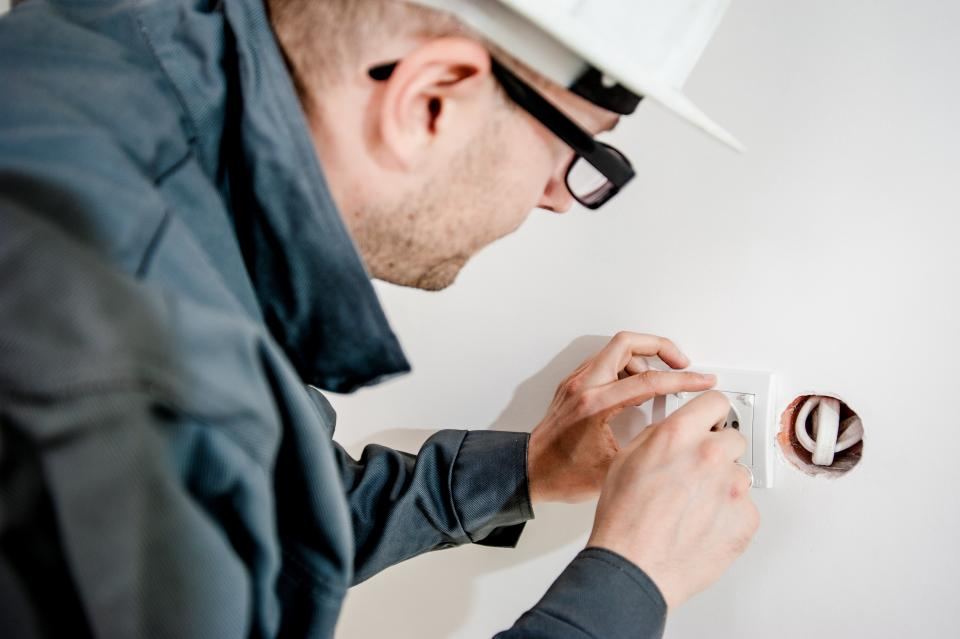 Een elektricien met helm schroeft een inbouwstopcontact vast in een witte muur.